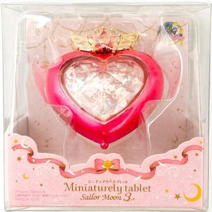 Könnte beinhalten: Ein pinkfarbenes herzförmiges Tablet mit facettiertem Mittelteil, goldener Krone und Flügeln. Die Verpackung enthält den Text "Miniaturely tablet Sailor Moon 3." und ist mit Bändern, Sternen und Monden verziert. Der Artikel befindet sich in einer transparenten Kunststoffhülle.