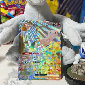 May include: A holographic Mew ex trading card featuring a pink Mew character, 180 HP, and Chinese text. The card displays a vibrant, multi-colored foil pattern with geometric shapes, set against a white plush toy background with blue accents.