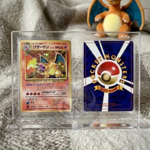 May include: A clear acrylic display case holding two Pokémon trading cards. The front card features a Charizard illustration with Japanese text. The back card displays the Pokémon logo. A Charizard plush toy sits in the background.