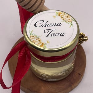 Könnte beinhalten: Ein kleines Glas Honig mit einem Deckel mit der Aufschrift "Chana Tova" und floralen Designs. Ein hölzerner Honiglöffel und ein rotes Band sind befestigt. Das Glas steht auf einem Holzuntersetzer.