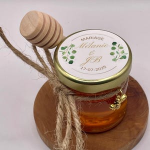 Può includere: Un piccolo barattolo di vetro pieno di miele, con coperchio dorato ed etichetta "Mariage Mélanie & JB 17-07-2025". Un cucchiaio di miele di legno e spago avvolgono il barattolo. Un piccolo ciondolo a forma di ape dorata pende dal barattolo.