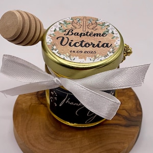 Può includere: Un piccolo barattolo di vetro con un coperchio dorato e un nastro bianco legato intorno. Il barattolo ha un'etichetta che dice "Baptême Victoria 14.09.2025". Il barattolo è appoggiato su una superficie di legno.