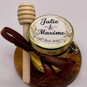 May include: A small glass jar filled with honey, decorated with a brown ribbon and a wooden honey dipper. The jar has a label that reads "Julie & Maxime 21 Juin 2025".