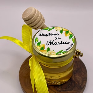 Può includere: Un piccolo barattolo di vetro pieno di miele, sormontato da un cucchiaio di miele in legno. Il coperchio presenta un'etichetta con la scritta "Baptême De Marius" e illustrazioni di limoni. Un nastro giallo è legato attorno al barattolo, su una base di legno.
