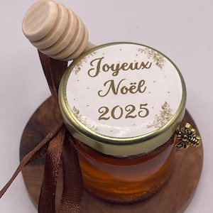 Peut inclure: Un petit pot de miel en verre, surmonté d'un couvercle avec l'inscription "Joyeux Noël 2025". Un bâtonnet à miel en bois et un ruban marron sont attachés. Le pot repose sur une base ronde en bois, avec un charm en forme d'abeille dorée.