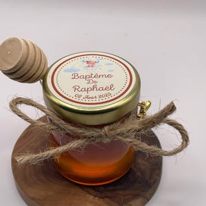 Può includere: Un piccolo barattolo di vetro pieno di miele, sormontato da un coperchio con la scritta "Baptême De Raphaël" e un'etichetta decorativa. Un cucchiaino di miele in legno poggia sul barattolo. Il barattolo è legato con spago e poggia su una base di legno.
