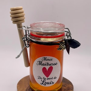 Può includere: Un piccolo barattolo di vetro pieno di miele, con un mestolo di legno per il miele attaccato al lato. Il barattolo ha un'etichetta che dice "Merci Maitresse" con un cuore rosso e "De la part de Louis".