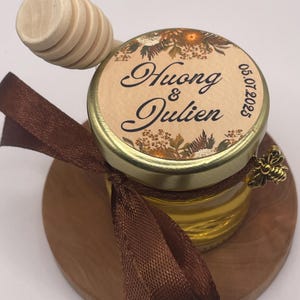 Può includere: Un piccolo barattolo di vetro pieno di miele, sormontato da un coperchio dorato e un cucchiaino di legno per il miele. Il coperchio presenta i nomi "Huong & Julien" e la data 05.07.2025. Un nastro marrone e un ciondolo a forma di ape decorano il barattolo, su una base di legno.
