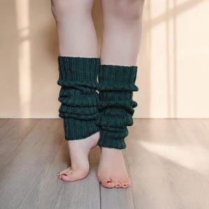Può includere: Un paio di scaldamuscoli verde bosco scuro con una texture a maglia a coste. Gli scaldamuscoli si estendono dalla parte inferiore del polpaccio alla caviglia, con un aspetto morbido e arricciato creato dal motivo a maglia verticale sulle gambe.