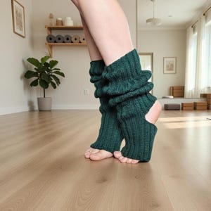 May include: Dark green knitted leg warmers with a ribbed texture. The leg warmers have a cutout for the heel and are worn on bare feet. The background is a light-colored wooden floor.