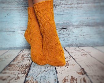 May include: A pair of bright orange knitted socks with a detailed cable knit pattern. The socks are displayed on a wooden surface with a blue and white painted background. The socks are a warm, cozy accessory.