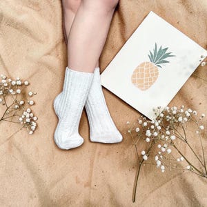 May include: A pair of white ribbed knit socks featuring a vertical textured pattern. The socks are positioned near a white rectangular card displaying a graphic illustration of a pineapple with a yellow body and green leaves, alongside sprigs of baby's breath.