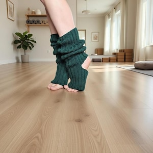 May include: A pair of dark green knitted leg warmers with a ribbed texture. The leg warmers have a cutout at the heel and are worn on bare feet. The background shows a yoga studio with a wooden floor and a small potted plant.