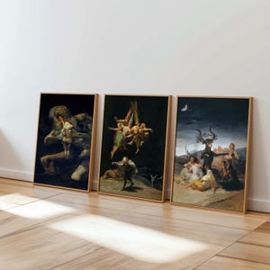May include: Three framed art prints featuring dark, classical paintings. The artworks depict mythological or allegorical scenes with figures in muted tones. Each piece is in a wooden frame, leaning against a white wall on a wooden floor.
