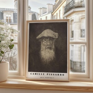May include: A framed print of a self-portrait by Camille Pissarro, displayed on a wooden windowsill. The black and white artwork depicts an elderly man with a long beard and hat. The print is in a light wood frame, with the artist's name and "Self Portrait" printed below.