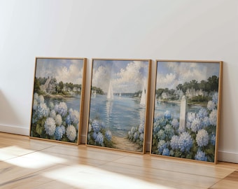 May include: A set of three framed landscape paintings featuring coastal scenes with blue and white hydrangea flowers in the foreground, sailboats on calm water, and houses along the shoreline under a cloudy sky, all encased in light wood frames.