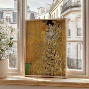 Könnte beinhalten: Ein gerahmter Druck eines Gemäldes von Gustav Klimt, das eine Frau in einem verzierten goldenen Kleid zeigt. Das Kunstwerk wird auf einer Fensterbank ausgestellt, mit Blick auf eine europäische Stadt im Hintergrund. Der Rahmen ist hellholzfarben.