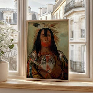 May include: A framed portrait of a Native American man with long black hair, a feather headdress, and red face paint. The artwork is displayed on a wooden windowsill, with a view of a European-style building in the background. The portrait is in a wooden frame.