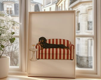 May include: A framed print featuring a dachshund dog lounging on a red and white striped sofa. A small side table holds a glass of wine. The artwork is set against a window with a view of a European city.