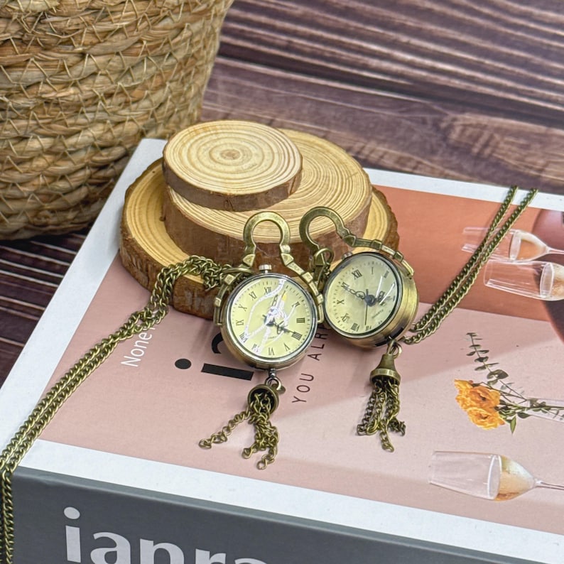May include: Two vintage-style pocket watch necklaces with bronze chains and tassels. The watch faces feature Roman numerals on a pale yellow background. The necklaces are displayed on a book. The word "None" is visible on the book.