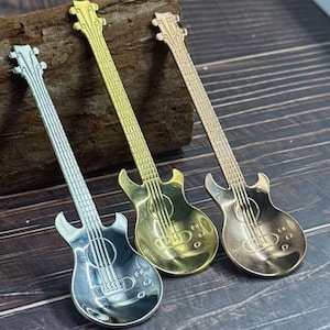 May include: Three decorative guitar-shaped spoons in silver, gold, and bronze tones. The spoons are arranged diagonally, with the silver spoon on the left, the gold in the center, and the bronze on the right. The spoons are resting on a wooden surface.