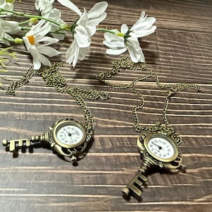 May include: Two antique-style key pendant watches with bronze-colored chains. The watch faces are white with black numerals and hands. The keys are detailed with intricate designs. White flowers are in the background.
