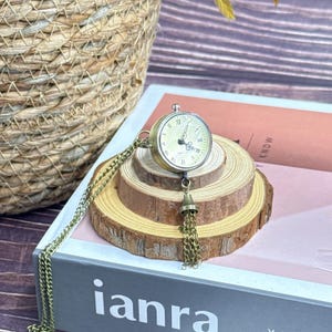 May include: A gold-coloured pocket watch pendant necklace. The watch face displays Roman numerals and fine hands. The necklace has a long chain and a decorative tassel. The watch sits on wooden slices, with the word "ianra" visible on a book.