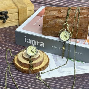 May include: Two antique-style pocket watches with bronze chains. One watch rests on a wooden slice, the other hangs from a wooden block. The watch faces are cream-coloured with black Roman numerals. The word "ianra" is visible on a book.