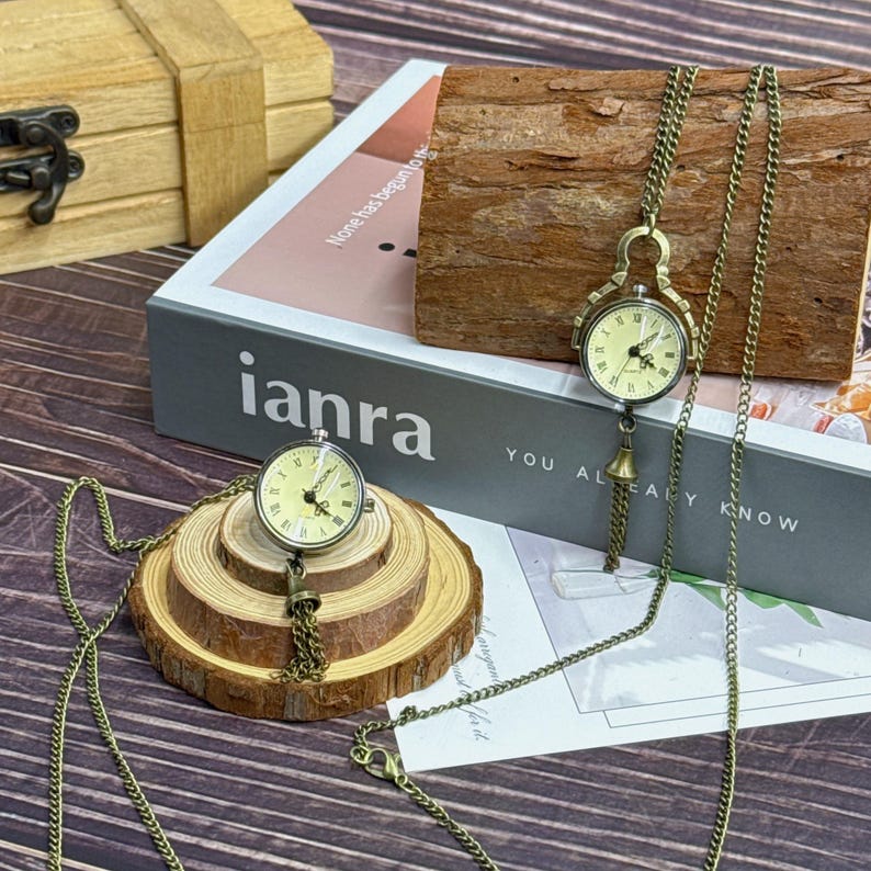 May include: Two antique-style pocket watches with bronze chains. One watch rests on a wooden slice, the other hangs from a wooden block. The watch faces are cream-coloured with black Roman numerals. The word "ianra" is visible on a book.