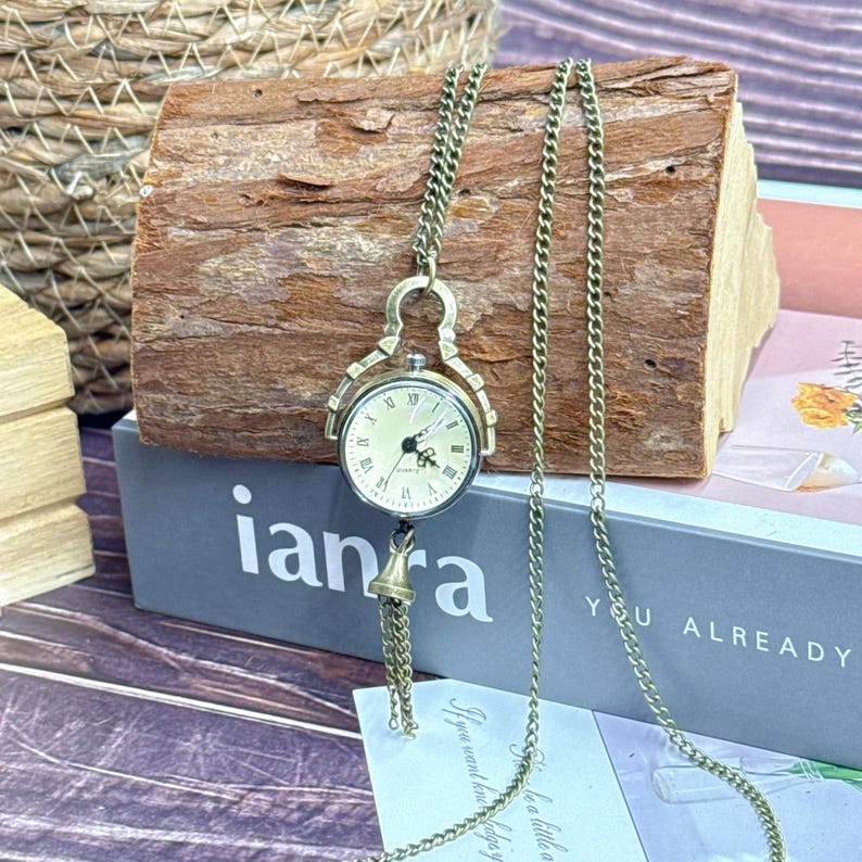 May include: Vintage-style pocket watch pendant necklace. The watch features a cream face with Roman numerals and a small bell charm. The chain and casing are bronze-coloured. The word "ianra" is visible in the image.