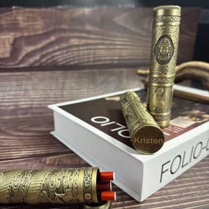 May include: Antique brass-colored cigarette holders with intricate engraved designs. One holder features a Buddha figure, another has a dragon motif, and a third is engraved with the name "Kristen". The holders are displayed on a book and wooden surface.