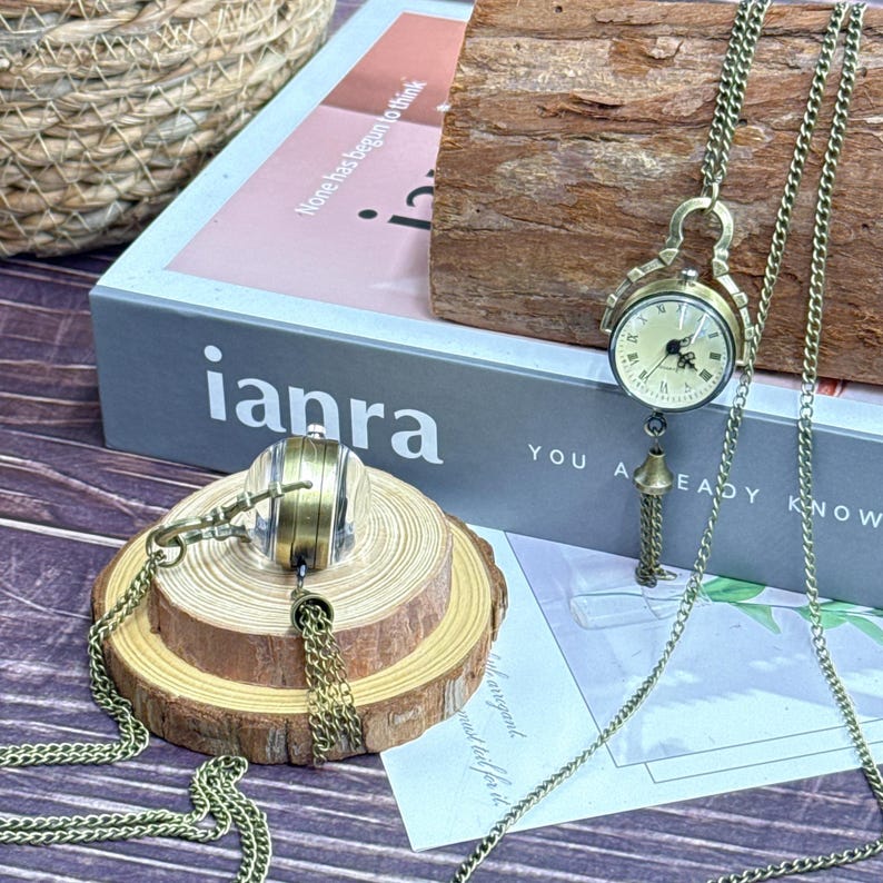 May include: Vintage brass-toned pocket watch pendant necklace with a cream clock face. The watch hangs from a long chain and has a decorative tassel. The watch is displayed on a wooden slice.