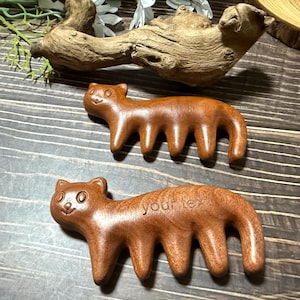 Può includere: Due pettini in legno intagliato marrone liscio a forma di animale, ciascuno con quattro denti. Il pettine inferiore presenta il testo inciso "your text.". Entrambi gli oggetti hanno orecchie arrotondate, tratti del viso semplici e finitura lucida.