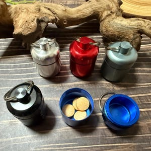 Pode incluir: Uma coleção de pequenos recipientes metálicos cilíndricos em prata, vermelho, cinza, preto e azul. Um recipiente está aberto, revelando vários comprimidos redondos e bege. Cada recipiente tem um porta-chaves prateado preso à tampa.