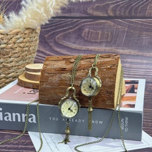 May include: Two antique-style pocket watch necklaces with bronze-coloured chains and watch cases. The watch faces are cream-coloured with black Roman numerals. Each watch has a decorative tassel. The text "YOU ALREADY NOW" is visible.
