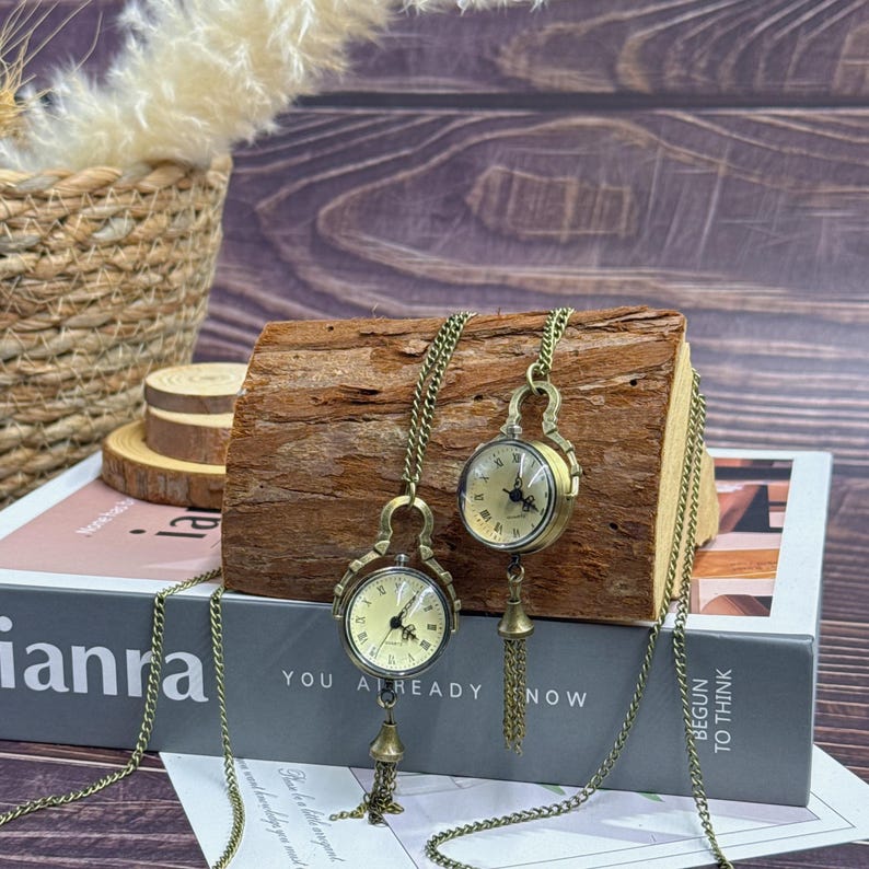 May include: Two antique-style pocket watch necklaces with bronze-coloured chains and watch cases. The watch faces are cream-coloured with black Roman numerals. Each watch has a decorative tassel. The text "YOU ALREADY NOW" is visible.