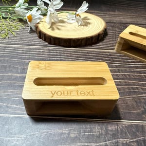 May include: A rectangular bamboo phone stand with a slot for the phone and the engraved text "your text". The stand is light brown and has a natural wood grain. A second stand is visible in the background.