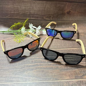 May include: Three pairs of sunglasses with black frames and light-coloured wooden arms. The lenses have a reflective, mirrored finish in shades of blue, orange, and grey. The sunglasses are arranged on a dark wooden surface.