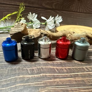 May include: Five small, cylindrical containers in blue, black, silver, red, and grey, each with a silver keyring. The black container has the text "YOUR TEXT" printed on it. The containers are arranged on a wooden surface.