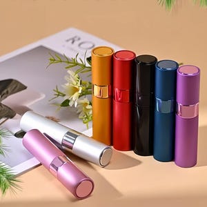 Op de afbeelding: Een collectie van zeven cilindrische navulbare parfumverstuivers in diverse kleuren, waaronder goud, rood, zwart, blauw, paars, zilver en roze. Elke verstuiver heeft een metallic afwerking, een centrale band en een platte dop aan beide zijden.