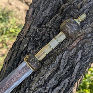 Könnte beinhalten: Ein römisches Gladius-Schwert mit silberner Klinge und dekorativem Griff. Der Griff hat einen dunkelbraunen, goldfarbenen Knauf und Parierstange mit einem elfenbeinfarbenen Griff. Die Klinge hat ein geätztes Design.