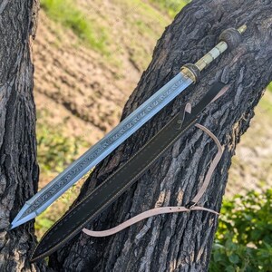 Könnte beinhalten: Ein römisches Gladius-Schwert mit detaillierter Klinge und kunstvollem Knauf, das auf einem Ast ruht. Das Schwert wird von einer schwarzen Lederscheide mit hellbraunem Riemen begleitet. Die Klinge weist geätzte Muster auf.