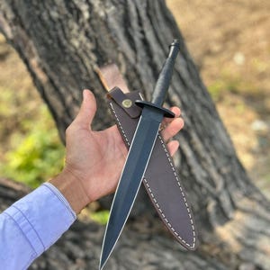 Puede incluir: Una daga negra con una hoja de doble filo y un mango negro. La daga se muestra con una funda de cuero marrón con costuras blancas y un cierre de botón a presión.