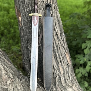 Op de afbeelding: Een zilveren zwaard met een bruine handgreep en gouden accenten, naast een zwarte schede. Het zwaardblad heeft een decoratief ontwerp en een rood embleem. Het zwaard wordt getoond tegen een boomstam.