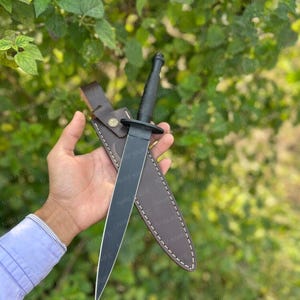 Peut inclure: Un poignard noir avec une lame droite et un manche noir est présenté dans un étui en cuir marron. Le poignard est tenu devant un fond de verdure verte. L'étui a des coutures blanches.