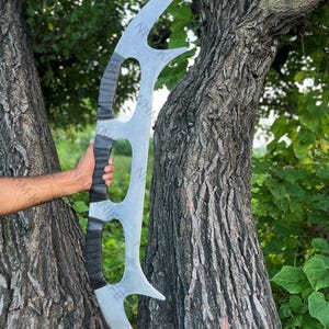 Könnte beinhalten: Eine große, silberfarbene, gebogene Klinge mit einem schwarzen Griff. Die Klinge hat eine einzigartige Form mit drei ovalen Aussparungen und zwei spitzen Enden. Der Griff scheint mit einem strukturierten Material umwickelt zu sein. Die Klinge wird vor einem Baum gehalten.