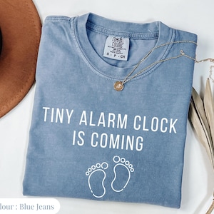 May include: A blue jeans colored Comfort Colors t-shirt with the text "TINY ALARM CLOCK IS COMING" in white. Below the text are two white footprints. A gold necklace with a pendant is on the shirt.