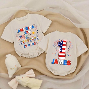 May include: Two off-white baby onesies. One features the text "AMERICA 250" with a flag, Statue of Liberty, and fireworks. The other has a large number "1" in red, white, and blue, with the name "JAMES" below.