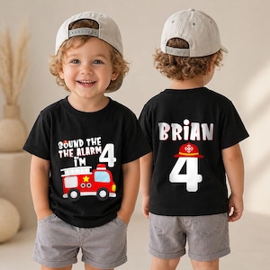 May include: A black t-shirt featuring a red fire truck graphic with the text "SOUND THE ALARM I'M 4" on the front and the name "BRIAN" above a red firefighter helmet and the number "4" on the back, paired with grey shorts and a beige baseball cap.