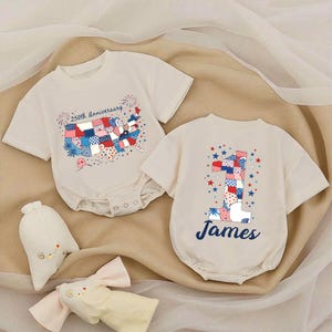 May include: Two off-white baby rompers. One features a map of the USA with a patchwork design and the text "250th Anniversary." The other has a large number "1" with a patchwork design and the name "James" in blue.